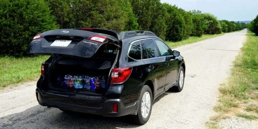 The image presents a scene featuring a black Subaru Outback parked on a country road with its trunk open.